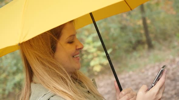 Slow motion of blond woman with yellow umbrella holding smart phone in the park 1080p HD footage - B