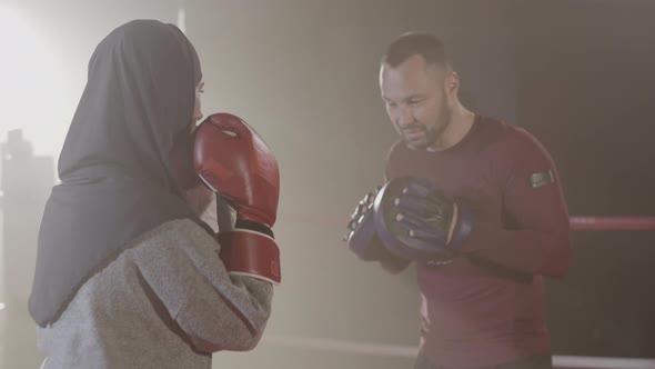 Back View of Young Woman in Hijab Punching Training Gloves of Coach in Backlight Fog. Confident