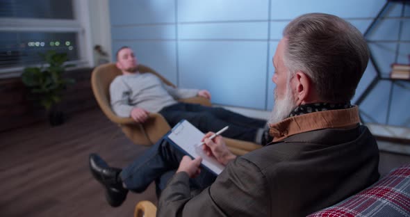 Professional Grayhaired Male Psychologist Listens to a Patient and Takes Notes on a Tablet During a