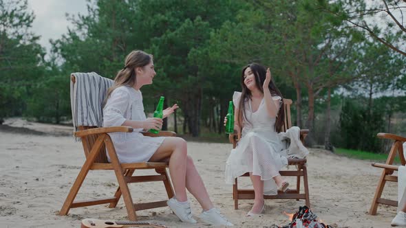 Young Women Talking and Cheering Bottles Sitting at the Campfire in the Woods