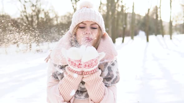 Young Woman in a Soft Pink Down Jacket White Hat Mittens and Scarf Blowing on the Snow on a