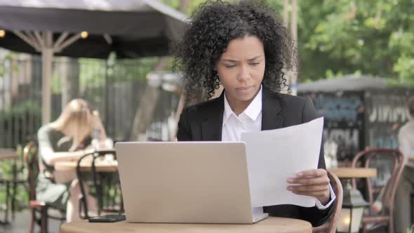 African Businesswoman Reading Contract and Using Laptop in Outdoor Cafe