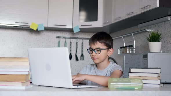 Boy with Glasses with Textbooks Doing Homework at School Using a Laptop at Home. Online Learning