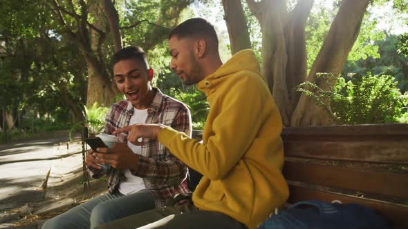 Two happy mixed race male friends sitting and using smartphone in park with backpacks