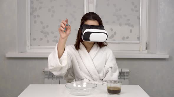 A Young Woman in a White Coat and VR Helmet Prepares Breakfast of Coffee and Cereals