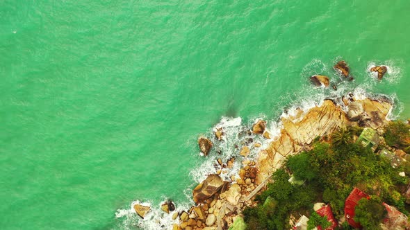 foamy waves of the emerald sea crashing on the rocky coast of Thailand. Colorful roofs on the edge o