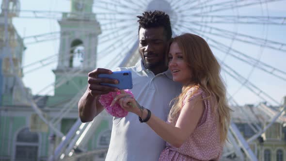 A Cheerful Couple Looks at the Smartphone Screen and Laughs in the Square