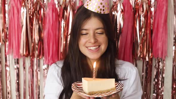 Happy woman in party hat making a wish and blowing candle on piece of birthday cake