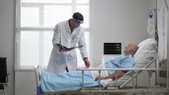 Young Doctor in Protective Mask Screen Talking to Senior Man Resting in Hospital Bed