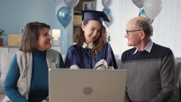 Family with Female Graduate in Hat and Gown are Listening to Congratulations Online Via Video