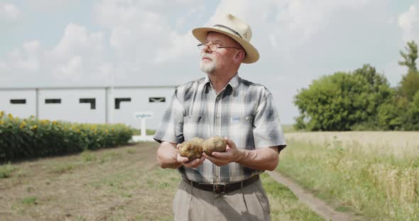 Farmer with Glasses in Hat Looks on Sun and Admires Potatoes in Hands at Field