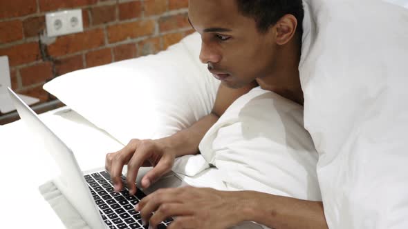 African Man Typing Email on Laptop, Lying on Stomach in Bed