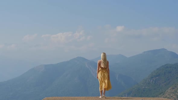 Young blonde woman in a yellow dress stands and looks at the mountains in Montenegro. Back view