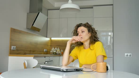 Tired Young Woman with Curly Hair and Yellow Shirt is Working From Home Using Her Laptop at the