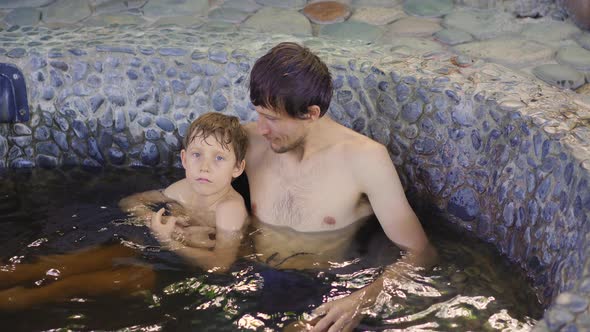 Young Man and His Son Relax in a Stone Bath Filled with Healing Herbal Infusions