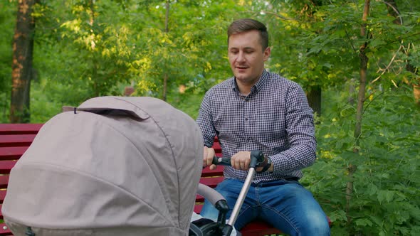 Happy Dad Rocking a Stroller with a Child Sitting in the Park. Dad Looks at the Baby in the Stroller