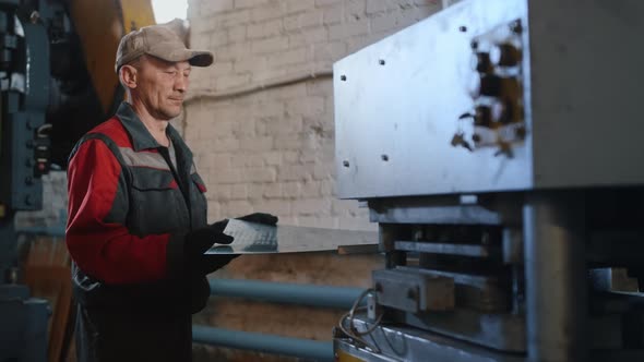 A Professional Caucasian Engineer in Overalls Processes a Sheet of Metal on a Machine