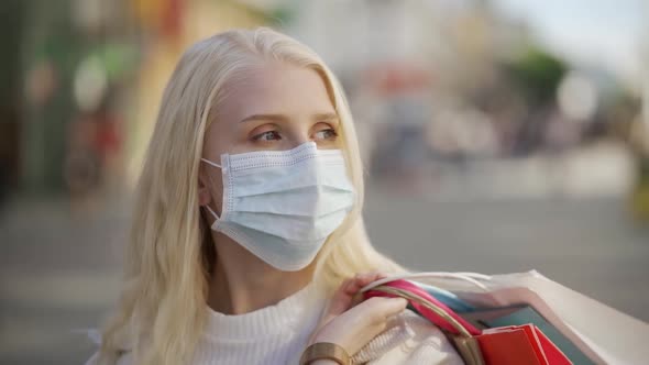 An Attractive Blonde Woman in a Medical Mask Walks Down the Street with Many Different Packages From