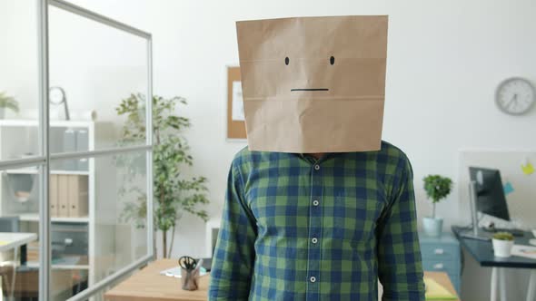 Portrait of Unhappy Guy Wearing Paper Bag with Emoji Standing at Work Shaking Head and Gesturing