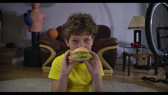 Portrait of Caucasian Boy Watching TV and Eating Hamburger. Cute Curly-haired Teenager with Junk