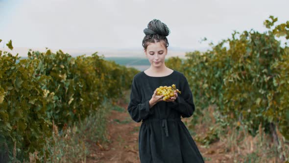 Young Woman with Dreadlocks Walking on the Path Among the Vineyard Showing White Grapes to the