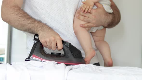 man is ironing a white shirt and holding a baby in arm,hand.multitasking husband is doing home stuff