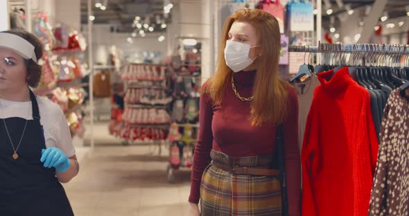 Shop Assistant in Safety Mask and Gloves Showing Sweater To Beautiful Girl in the Department Store