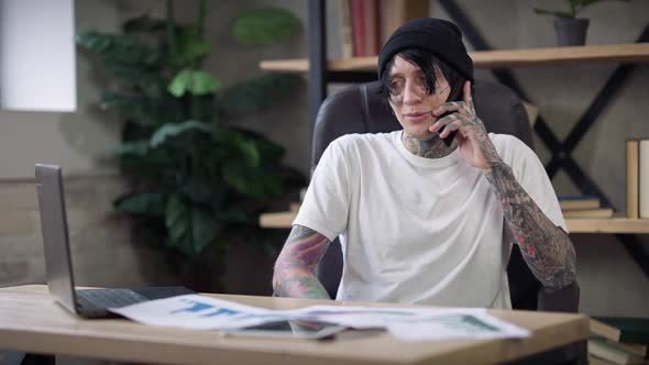 Cheerful Tattooed Man in White Tshirt and Black Hat Talking on Phone Sitting in Home Office on