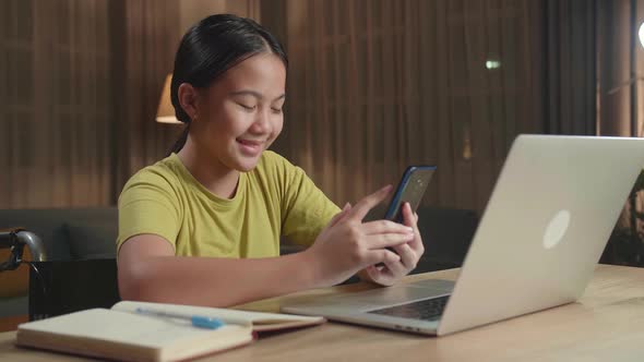 Asian Kid Girl Sitting In A Wheelchair While Using Laptop Computer And Use Mobile Phone At Home alt