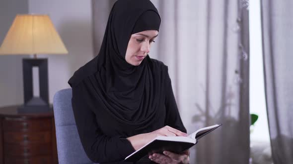 Portrait of Intelligent Muslim Woman in Hijab Reading Book at Home. Concentrated Young Woman Making