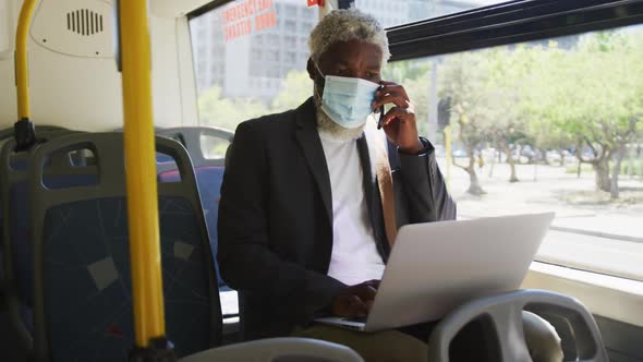 African american senior man wearing face mask talking on smartphone and using laptop while sitting i