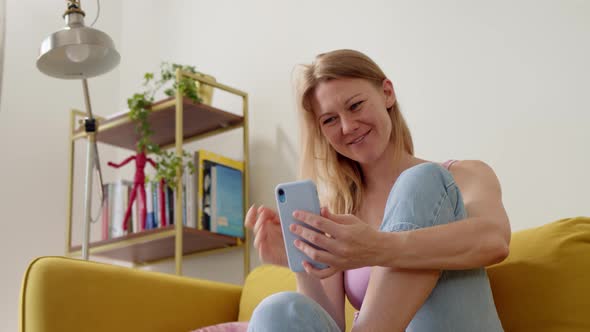 Happy Young Cheerful Blonde Woman in Pink Top Sits on Couch at Home and Video Call on Blue Phone