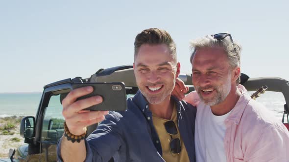 Happy caucasian gay male couple taking selfies and smiling by car on sunny day at the beach