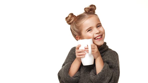 Small Beautiful Caucasian Kid Girl in Cozy Sweater Drinking From Cup on White Background and Smiling