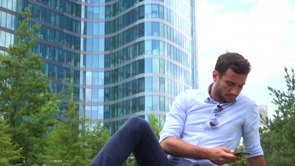 A Young Handsome Businessman Sits on Grass in a Park and Works on a Smartphone - Closeup