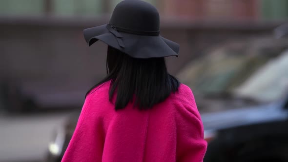 a Brunette in a Black Hat and a Bright Pink Coat Stands Against the Background of a Blurred City