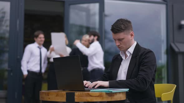 Serious Busy Young Employee in Suit Sitting on Office Terrace Messaging Online on Laptop with