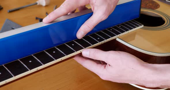 Close up hands of a luthier sanding and leveling the frets on an acoustic guitar neck fretboard on a