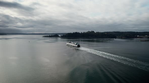 Following behind a large commuter ferry on dark calm water, gloomy clouds reflect, aerial