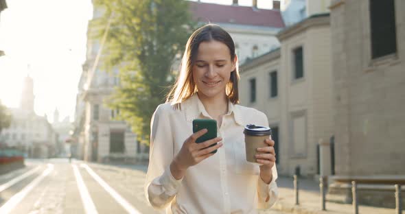 Attractive Young Woman Using Smartphone and Holding Coffee Cup While Standing in Sunlights