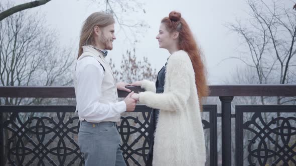 Side View of Happy Caucasian Man Standing at Bridge and Holding Hand of His Lovely Girlfriend