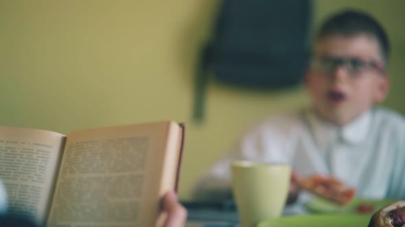 Schoolboy Reads Books Opposite Friend Eating Pizza at Table