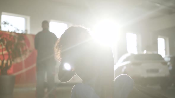 Beautiful Young Caucasian Woman Sitting in Sunrays in Auto Repair Shop and Smiling. Gorgeous Female