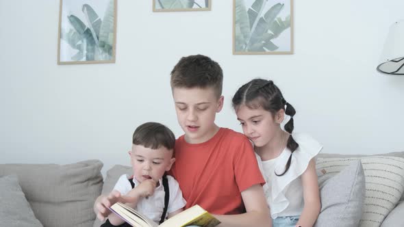 Three Children of Different Ages Read a Book Sitting on the Couch in the Living Room
