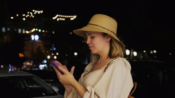 Handheld Shot of a Woman Using a Smartphone While Standing in the Middle of a Night City Street