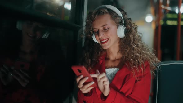 A Smiling Curly Woman in Headphones is Listening Music and Using Smartphone or Reading Messages in