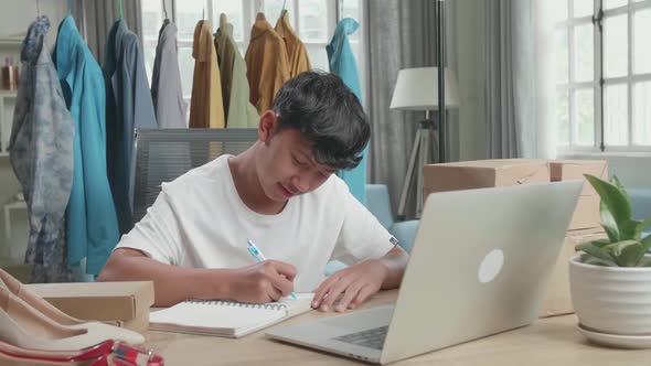 Asian Man Online Seller Looking At Computer Screen And Writing In The Notebook While Selling Clothes