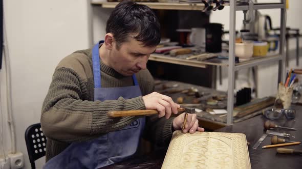 A Craftsman in Blue Overalls Carving Wooden Ornament on Backgammon Sitting in His Working Place