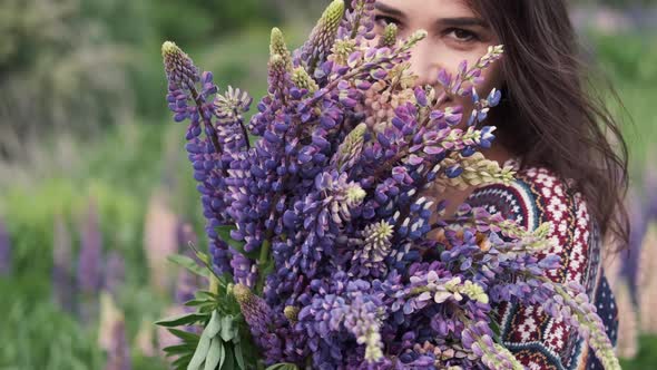 Young Beautiful Smiling Woman with a Bouquet Walks Among the Flowers of Purple Lupins