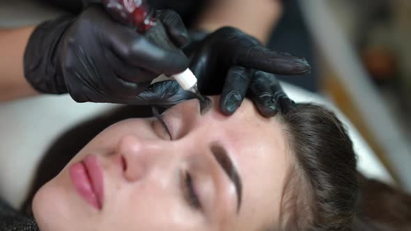 Face of Young Gorgeous Woman with Beautician Hands Tattooing Second Eyebrow with Microblading Pen